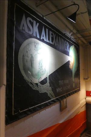 A mural painted on a white brick wall shows a face with bright lights coming out of the eyes casting a glow over a glass ball. Inside the ball are images of people engaged in various activities. The face is wrapped in a green question mark. The text says Ask Alexander The Man Who Knows.